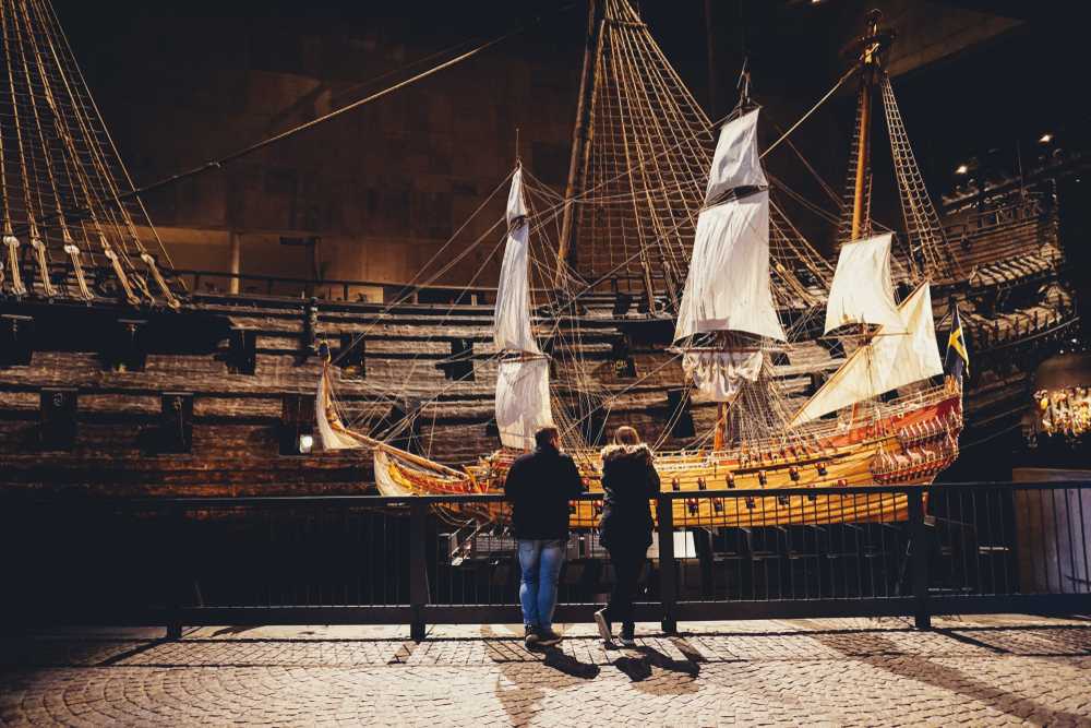 Two people lean on a black railing that separates them from a huge wooden ship on display in what looks like a naval museum.