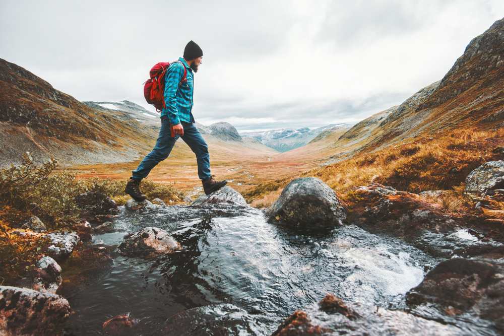A man in a cap and plaid shirt crosses a river in a natural wilderness where there are large stones.