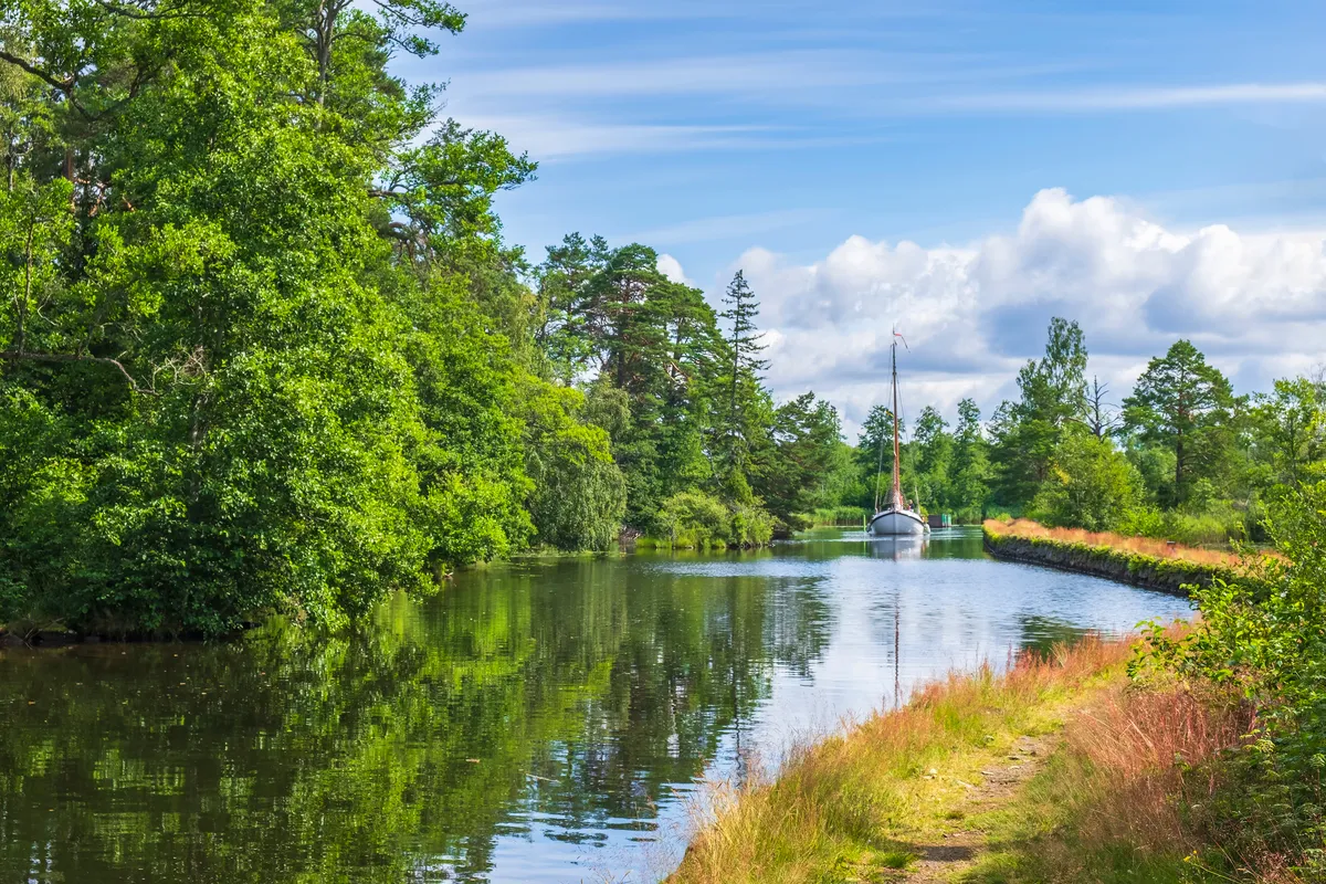Tree-lined stretch of the Göta Canal with a sailboat approaching, calm reflections, and a grassy towpath under a partly cloudy sky.