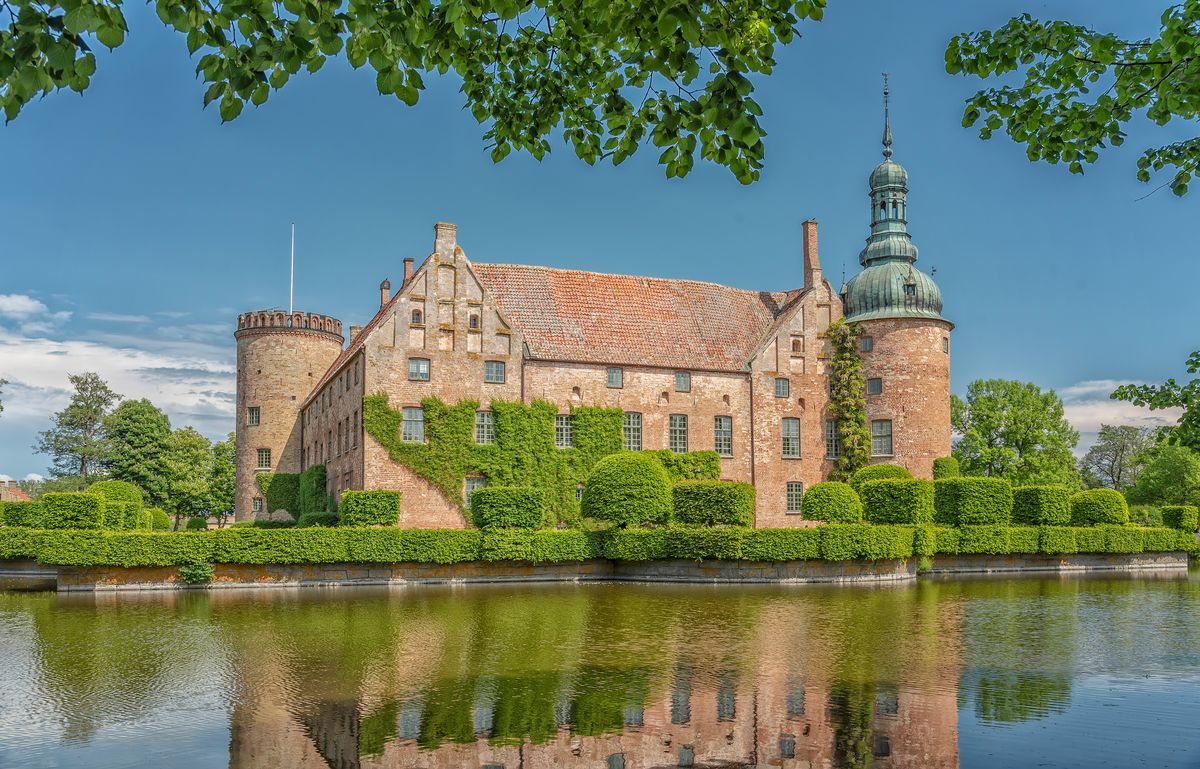 A beautiful postcard of a salmon-colored mansion located in front of a calm body of water that reflects its facade on a clear day, surrounded by green