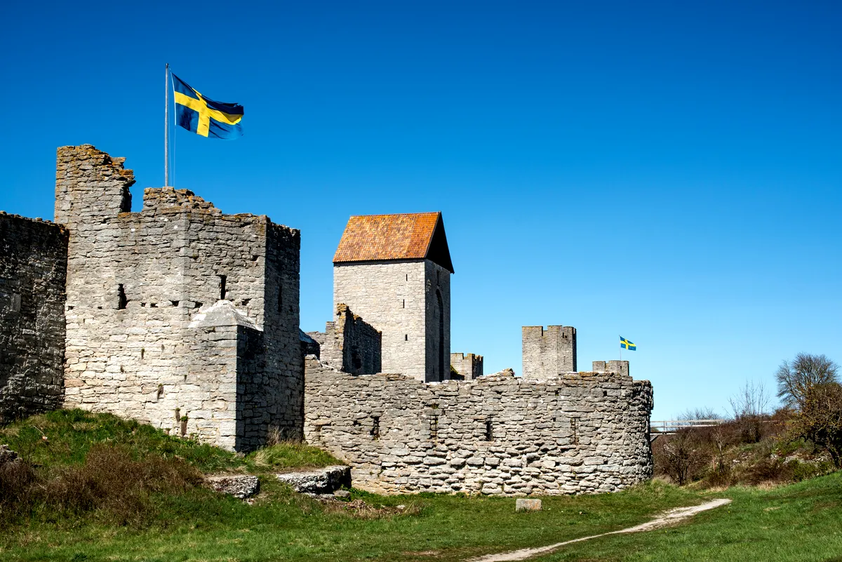 Medieval stone fortress walls and tower with Swedish flags under a clear blue sky, grassy path in the foreground.