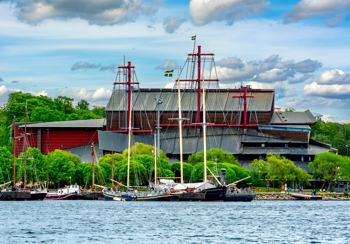 Tree-lined stretch of the Göta Canal with a sailboat approaching, calm reflections, and a grassy towpath under a partly cloudy sky.