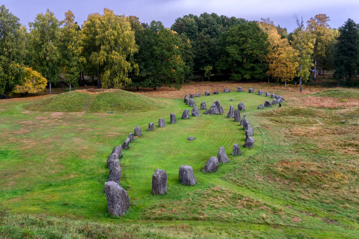 Eine Luftaufnahme des Anundshög-Grabhügels und der Steinschiffformation in Västerås, Schweden, umgeben von üppigem Grün und Bäumen mit Herbstlaub.