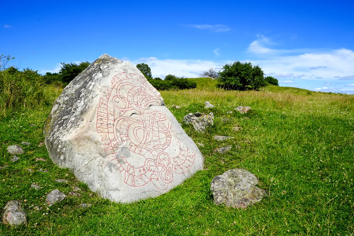 A Viking runestone at Birka, Sweden, standing in a grassy field under a clear blue sky, with intricate carvings and Norse inscriptions visible on its surface.