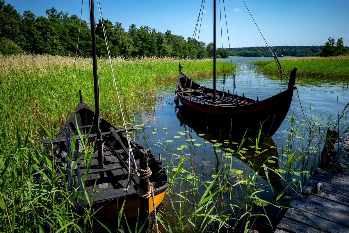 Two traditional Viking ships anchored in the serene waters of Birka, Sweden, surrounded by lush greenery and tall reeds, offering a glimpse into the rich maritime history of the Viking era.