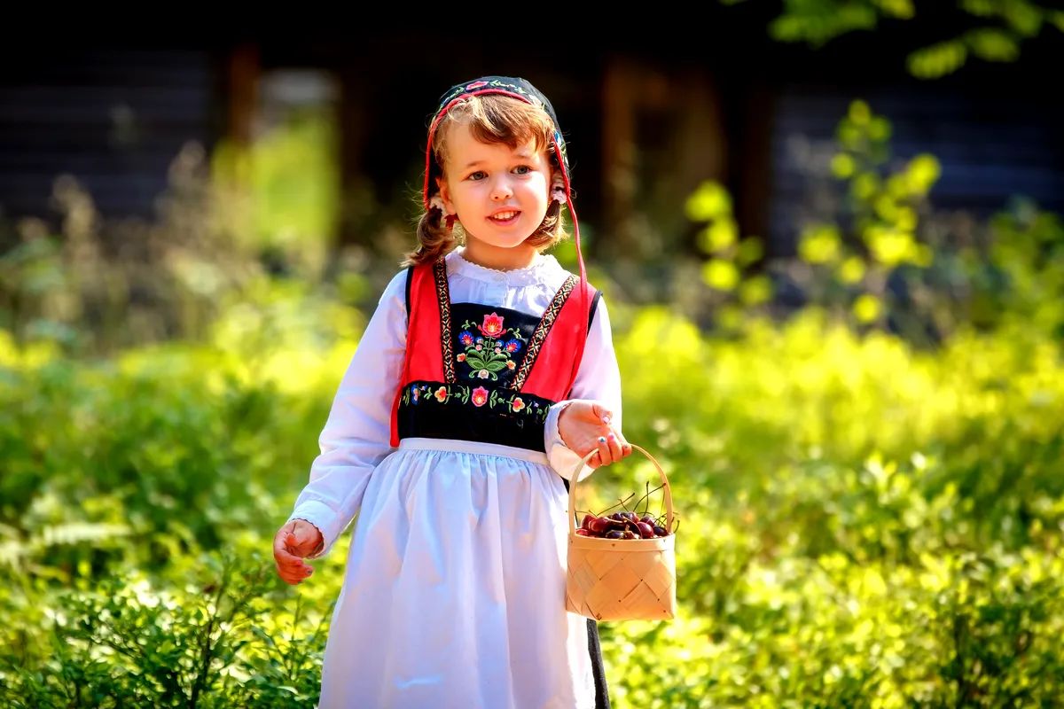 Young Girl in Traditional Swedish Folk Costume Holding a Basket of Berries A young girl wearing traditional Swedish folk clothing stands outdoors, holding a small basket filled with berries. She is dressed in a colorful outfit with intricate floral embroidery on the bodice and a headscarf that matches her dress. The sunlight filters through the trees, illuminating her with a warm glow, creating a picturesque and charming scene of Swedish cultural heritage. The background features lush greenery, enhancing the peaceful and idyllic atmosphere.