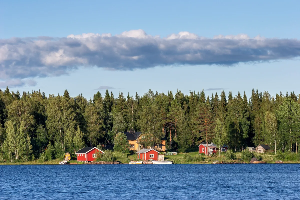 Traditionella svenska stugor vid en sjö i Luleå skärgård Naturskön utsikt över traditionella svenska röda trästugor inbäddade bland täta gröna skogar längs stranden av en lugn blå sjö. Stugorna är omgivna av frodiga träd, och några små båtar ligger förtöjda nära strandlinjen. Himlen är klar med en Få spridda moln skapar en lugn och idyllisk atmosfär som är typisk för den svenska landsbygden. width=