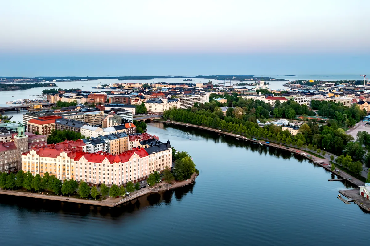An aerial view of Helsinki, Finland, showcasing the city's unique blend of historic and modern architecture. The image captures the vibrant cityscape with colorful buildings, particularly the red-roofed structures in the foreground, surrounded by lush green trees and waterways. The city stretches out towards the horizon, with the Baltic Sea and small islands visible in the distance, creating a picturesque coastal scene under a clear, soft blue sky.