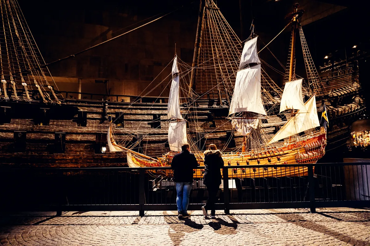 A dimly lit museum exhibit featuring a large, historic wooden ship with tall masts and sails, reminiscent of the 17th century. Two visitors, a man and a woman, stand close together, observing the intricate details of a smaller, brightly colored ship model displayed in the foreground. The scene evokes a sense of awe and fascination with maritime history, highlighting the rich cultural heritage preserved in the museum.