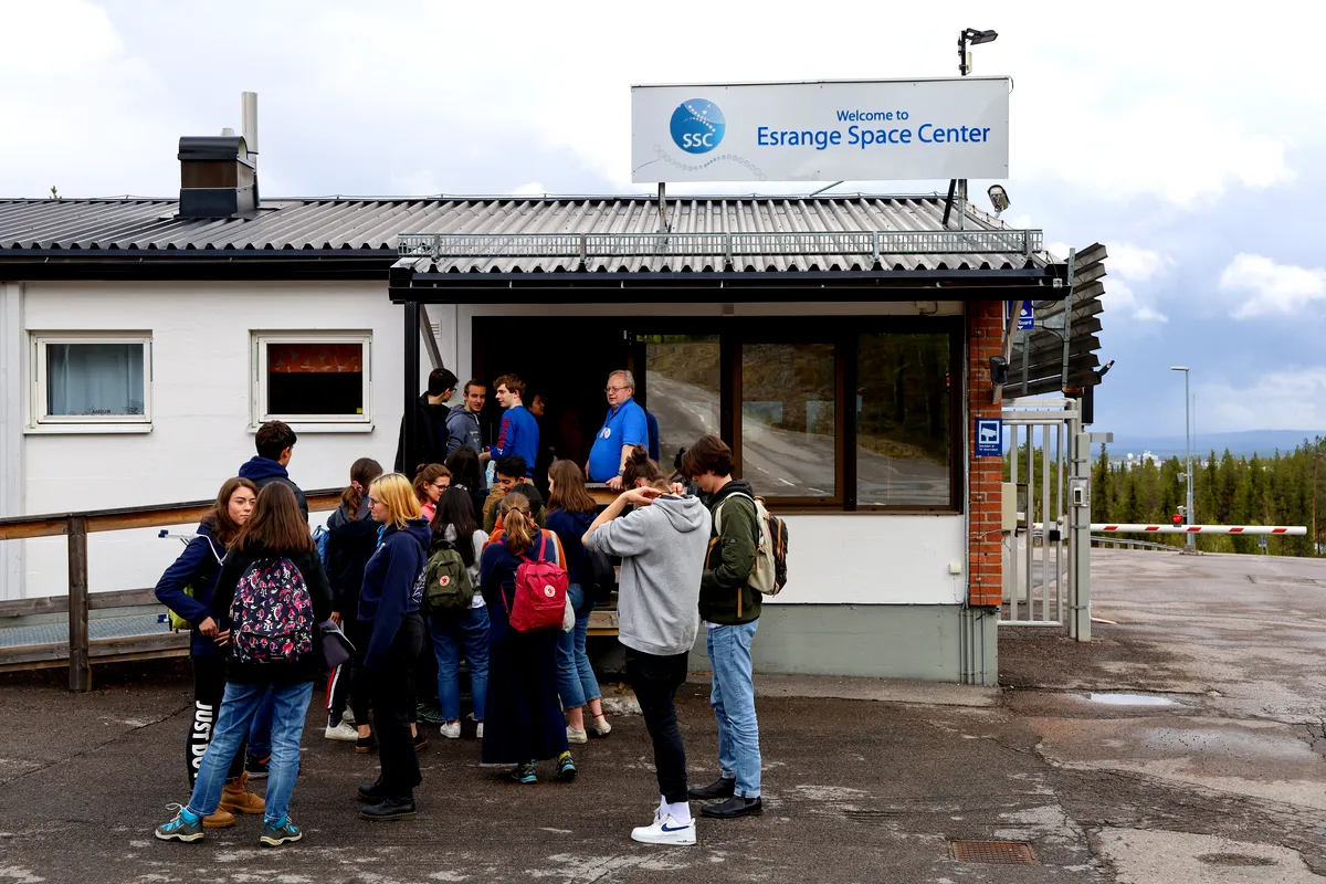 Group of tourists gathered at the entrance of Esrange Space Center in Sweden, preparing for a guided tour, with a signboard and cloudy skies in the background.