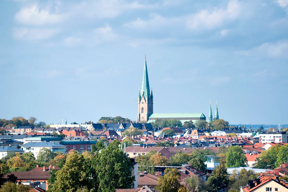 Panoramic View of Linköping: The Iconic Cathedral Rising Above the Cityscape A panoramic view of Linköping, Sweden, with its iconic cathedral dominating the skyline. The cathedral's tall, green spire rises above the surrounding buildings, which are a mix of traditional and modern architecture. The city's rooftops, covered in red and brown tiles, blend harmoniously with the lush green trees scattered throughout the urban landscape. The bright, clear sky enhances the serene and picturesque atmosphere of this historical city.