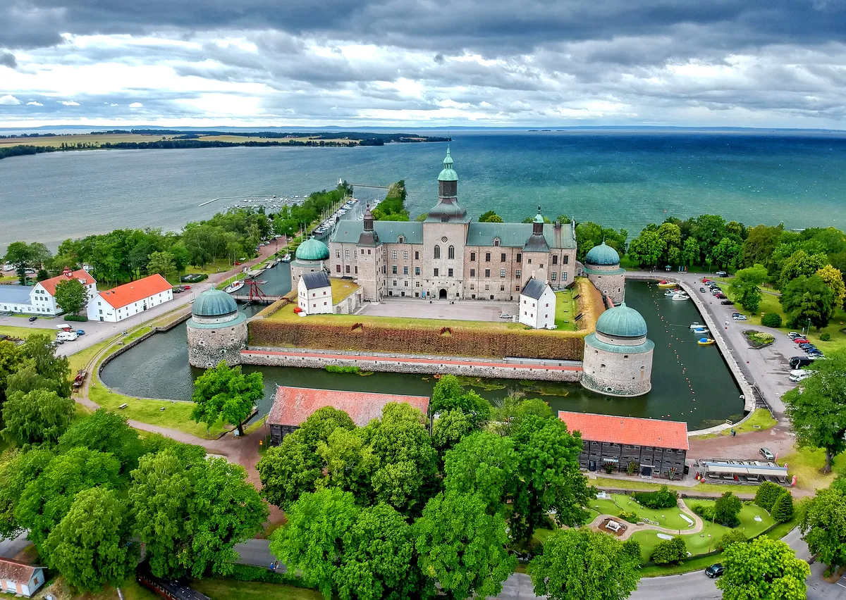 Aerial View of Vadstena Castle: Renaissance Architecture by Lake Vättern Aerial view of Vadstena Castle, an iconic Renaissance fortress in Sweden, surrounded by lush green trees and bordered by the calm waters of Lake Vättern. The castle, with its distinct turrets and domed roofs, stands majestically within a moat, highlighting its historic architectural style. The bright, green landscape and the vibrant blue of the lake under a partly cloudy sky create a picturesque and serene setting, reflecting the castle’s rich historical significance.