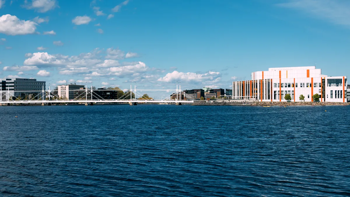 Stadsvy och bro över en lugn sjö En modern stadsvy sedd över en lugn sjö under en klarblå himmel. Horisonten har samtida byggnader, inklusive en stor vit och orange byggnad, samt en elegant vit bro som korsar vattnet. Reflektionerna på vattnet bidrar till den fridfulla atmosfären. Detta urbana område är omgivet av stilla vatten och öppna skyar, vilket ger en rofylld utsikt.