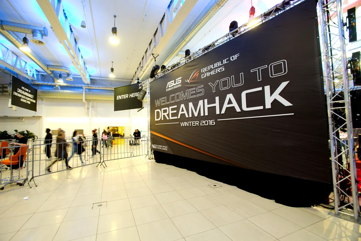 Ingång till DreamHack Winter 2016 En stor banderoll inuti en modern, väl upplyst lokal välkomnar deltagare till “DreamHack Winter 2016” sponsrat av ASUS Republic of Gamers. Människor syns gå genom ett avdelat område på väg mot evenemanget. Lokalen har ljusa lampor, rena kakelgolv och metalliska takdetaljer. Evenemangsutrymmet ser organiserat ut och redo för ett storskaligt gaming- eller teknikevent.