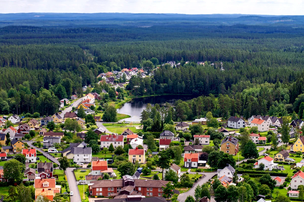 Flygfoto över en svensk stad omgiven av skog Flygfoto över en liten stad omgiven av frodig grön skog. Staden består av färgglada hus med trädgårdar, prydligt ordnade längs slingrande gator. I bakgrunden syns en fridfull sjö omgiven av täta tallar. Horisonten sträcker sig ut i en vidsträckt skog, vilket ger en känsla av lugn och kontakt med naturen.