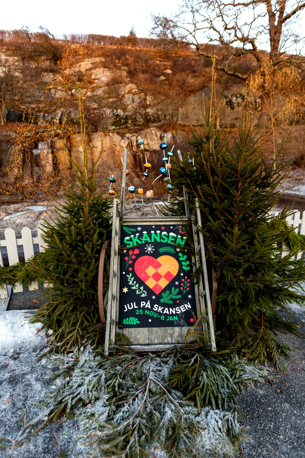 Ein festliches, weihnachtlich gestaltetes Schild in Skansen, aufgestellt in einer natürlichen Umgebung im Freien mit Kiefern und Weihnachtsdekorationen. Das Schild zeigt ein farbenfrohes gewebtes Herzdesign mit den Worten „Skansen“ oben und dem Text „Weihnachten in Skansen, 25. November – 8. Januar“ unten. Das Schild ist von winterlichem Grün und einer frostigen Landschaft umgeben und vermittelt eine gemütliche, weihnachtliche Atmosphäre.