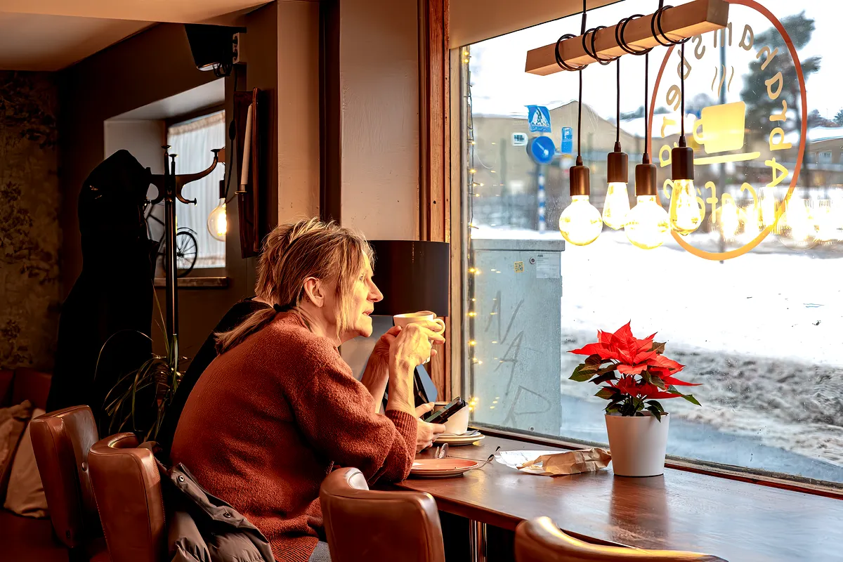 Eine Frau sitzt am Fenster eines gemütlichen Cafés in Stockholm, Schweden. Sie hält eine warme Tasse Kaffee in den Händen und blickt mit einem entspannten Gesichtsausdruck nach draußen. Das Café hat eine warme, einladende Atmosphäre mit hängenden Glühbirnen und einem Weihnachtsstern auf dem Tisch. Vor dem Fenster ist Schnee zu sehen, was zur gemütlichen Atmosphäre der Szene beiträgt.