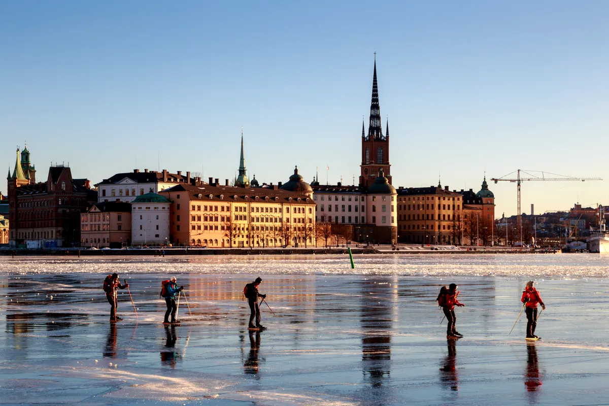 Eine Gruppe von Menschen läuft auf einem zugefrorenen See in Stockholm, Schweden, Schlittschuh. Die Schlittschuhläufer in Winterkleidung gleiten über die glänzende Oberfläche des Eises und halten mit Skistöcken das Gleichgewicht. Im Hintergrund sind die historischen Gebäude und Kirchtürme der Stadt zu sehen, getaucht in warmes Sonnenlicht vor einem klaren blauen Himmel. Die Szene spiegelt die Winteraktivitäten wider, die die Einheimischen in der Stadt während der kälteren Monate genießen.