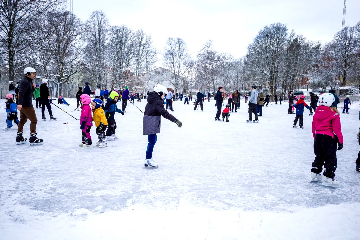 Eine lebhafte Winterszene in Stockholm, wo Kinder und Erwachsene auf einer Eisbahn im Freien Schlittschuh laufen. Alle sind in farbenfrohe Winterkleidung eingepackt, und die Kinder tragen aus Sicherheitsgründen Helme. Der Boden ist mit Schnee bedeckt und die umliegenden Bäume sind kahl, was zur winterlichen Atmosphäre beiträgt. Man sieht Familien und Freunde, die beim Schlittschuhlaufen lachen und Spaß haben.