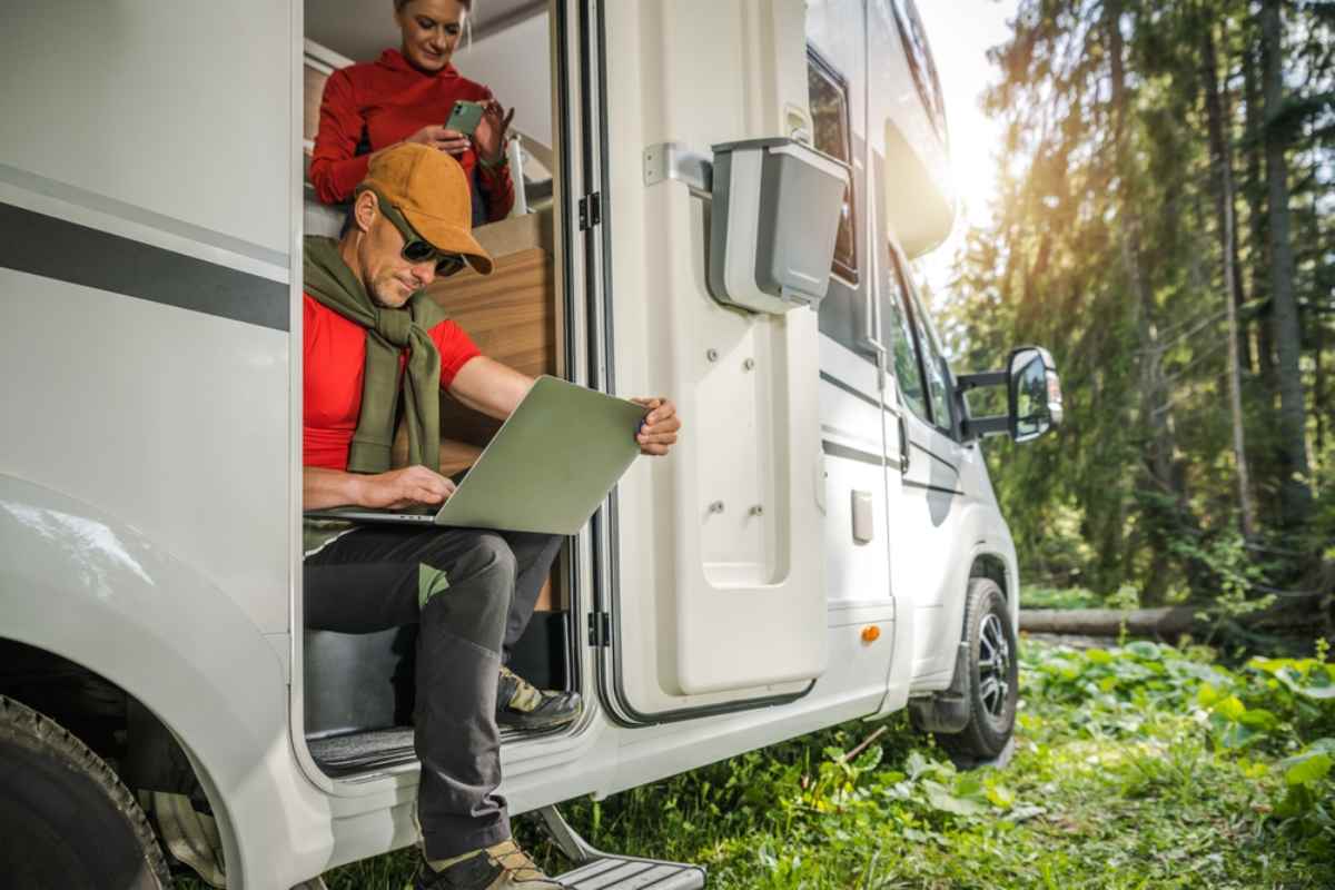 A couple, the capped man sitting almost outside their parked camper and the woman inside, peruse the screens of their various electronic devices.